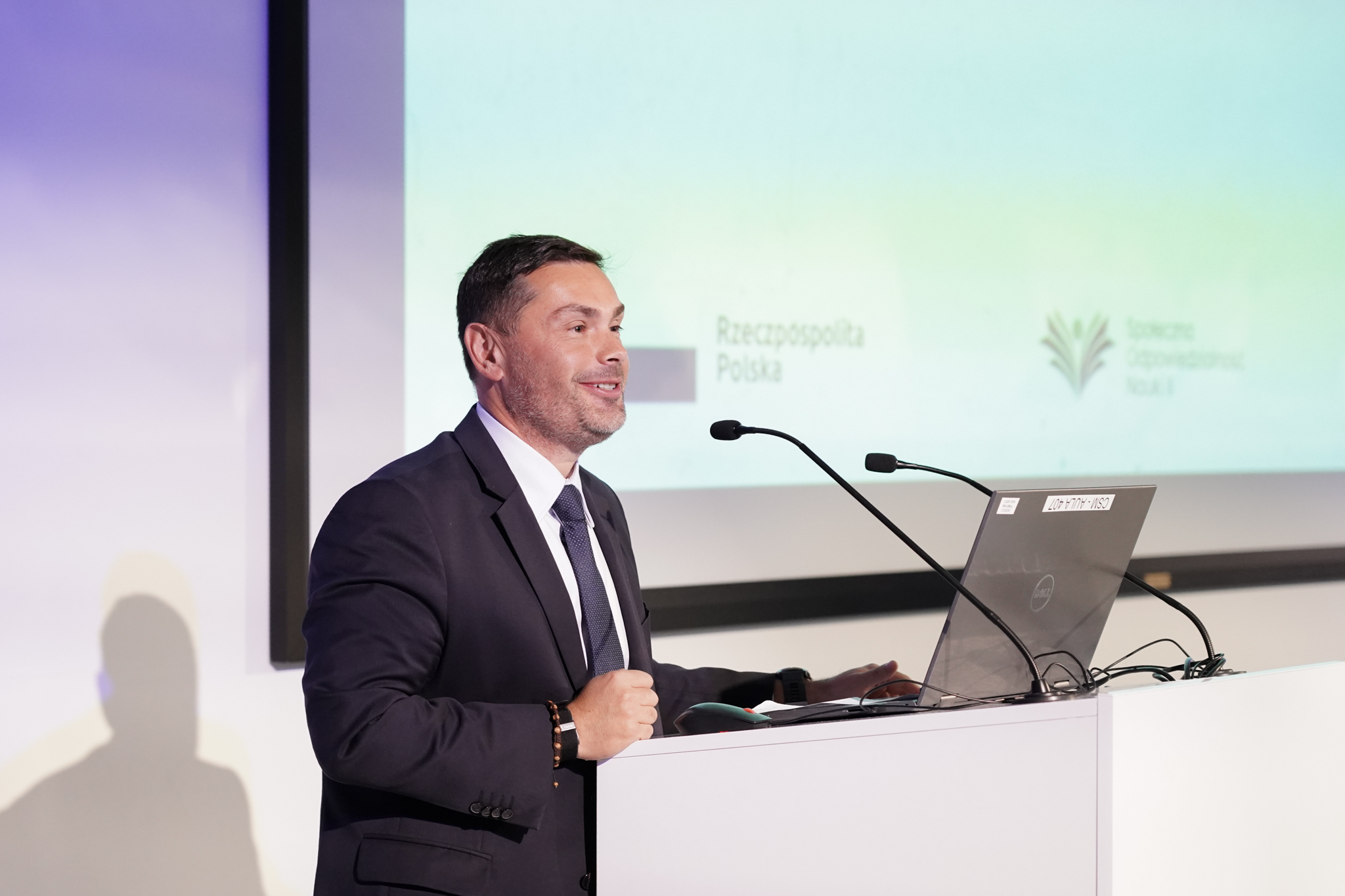 Elegancko ubrany i uśmiechnięty człowiek w średnim wieku, przed m&oacute;wnicą, dwoma mikrofonami i laptopem, z tyłu wyświetlana prezentacja